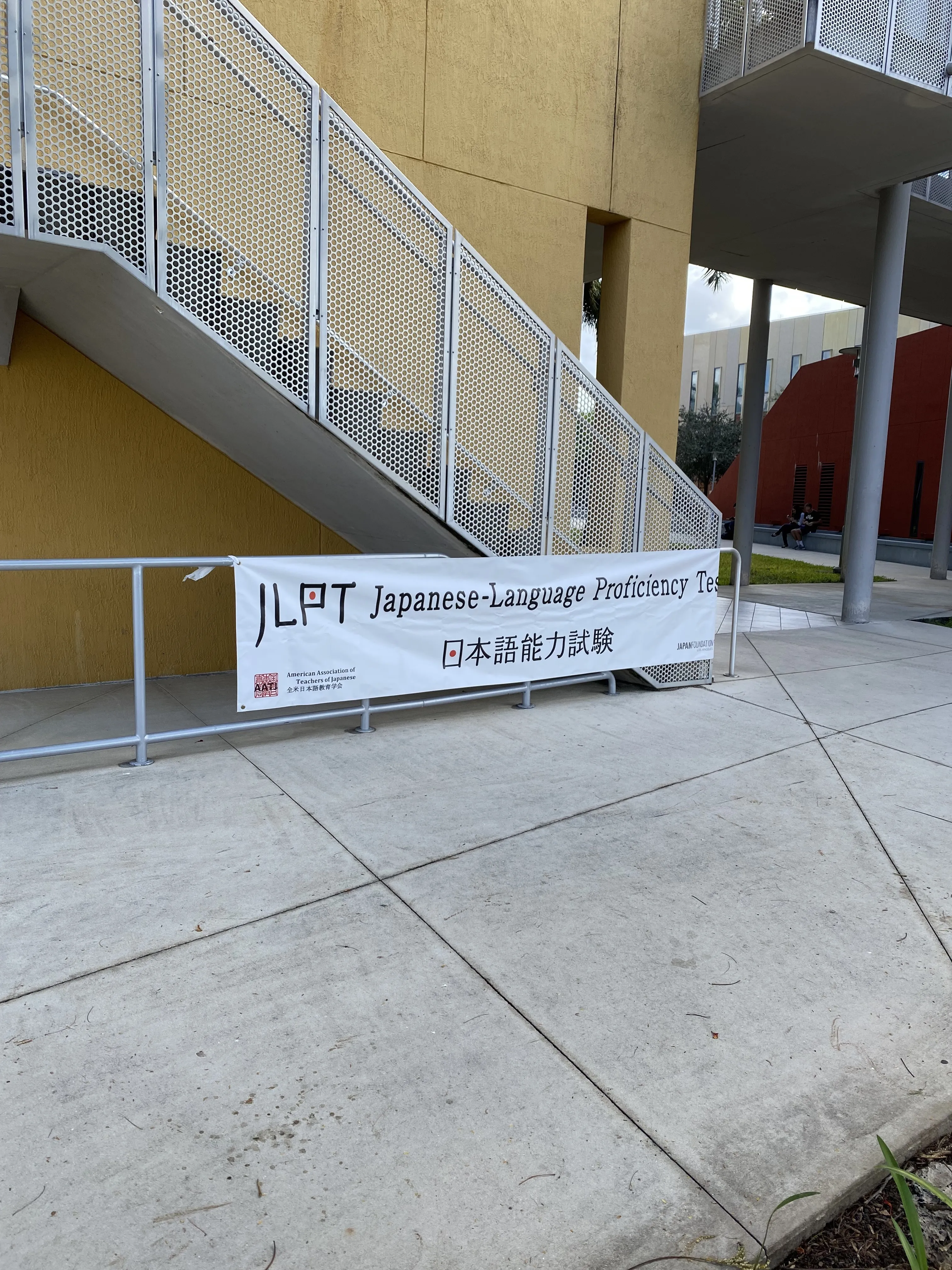 The JLPT testing-site banner zip-tied to a stair railing outside the FIU College of Business Complex, reading "JLPT Japanese-Language Proficiency Test" with the Japanese 日本語能力試験 below it.