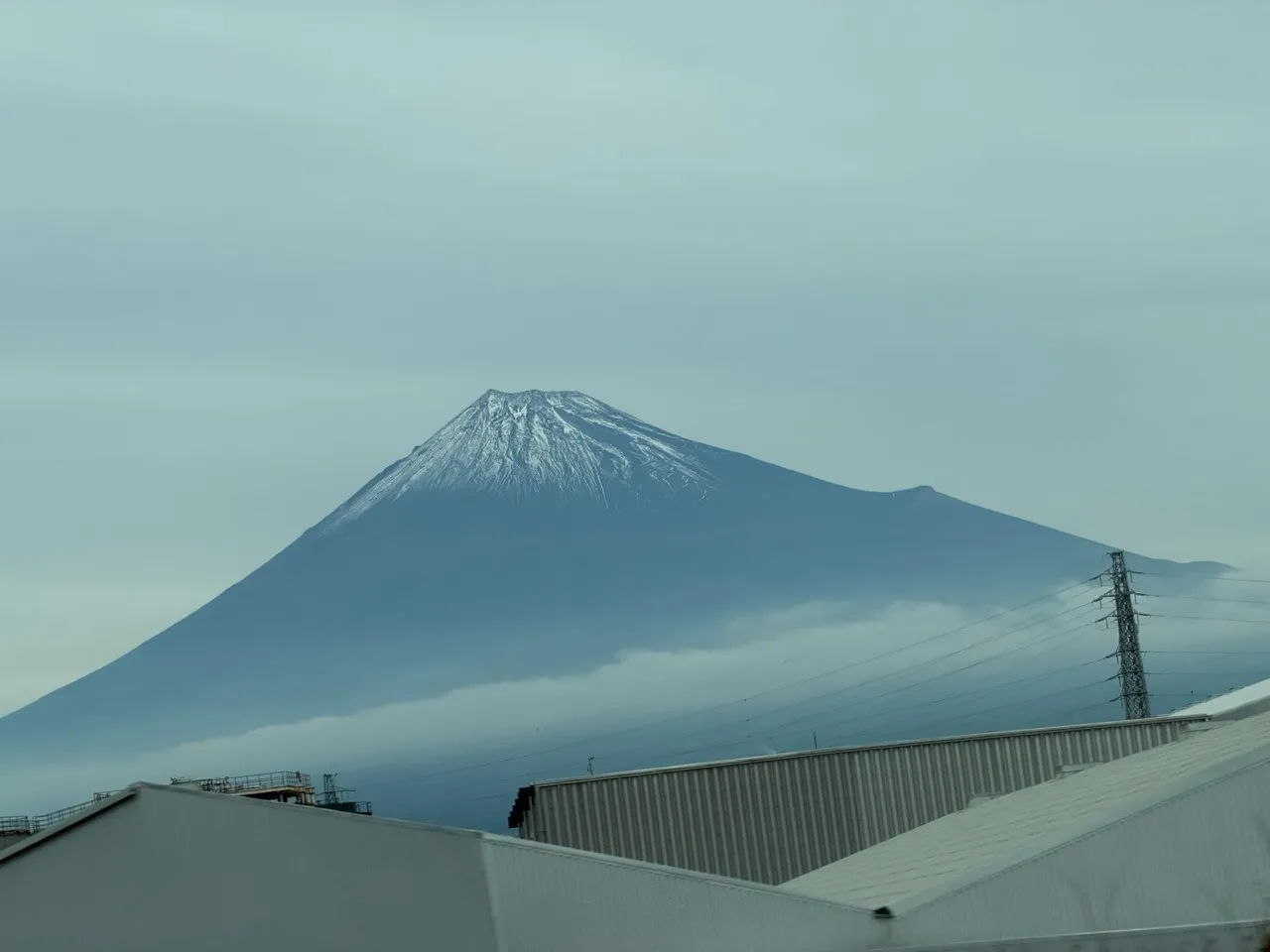 Snow-capped Mount Fuji rising above industrial buildings, shot from a moving train