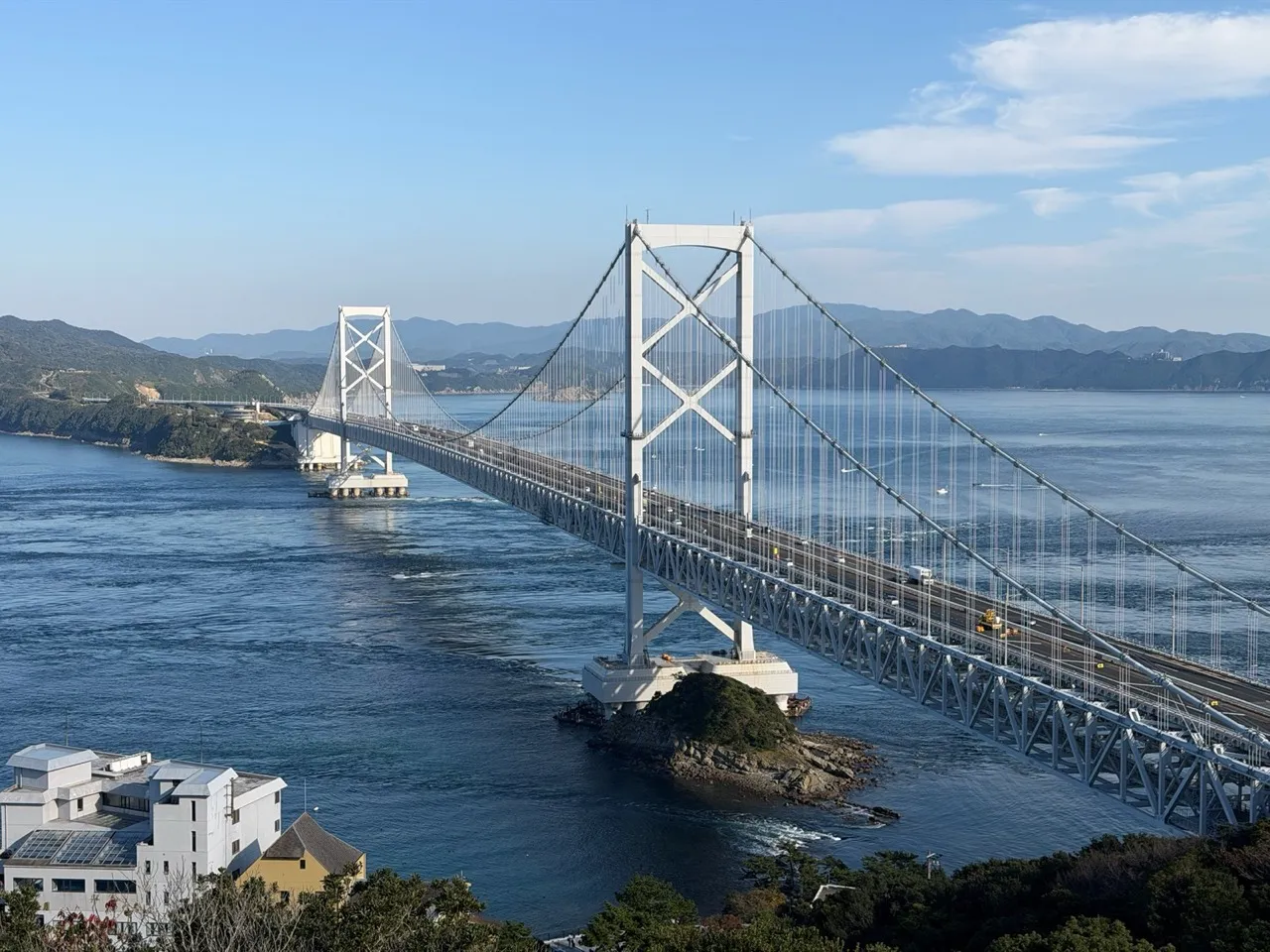 A large white suspension bridge spanning a blue ocean strait between forested headlands