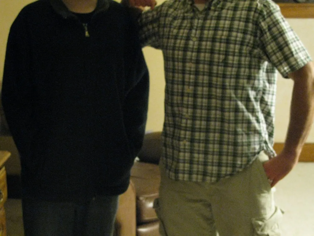 Todd and Craig standing side by side, photographed from the neck down so neither face is in frame: Todd in a dark top on the left, the much taller Craig with his arm around him in a plaid shirt on the right.