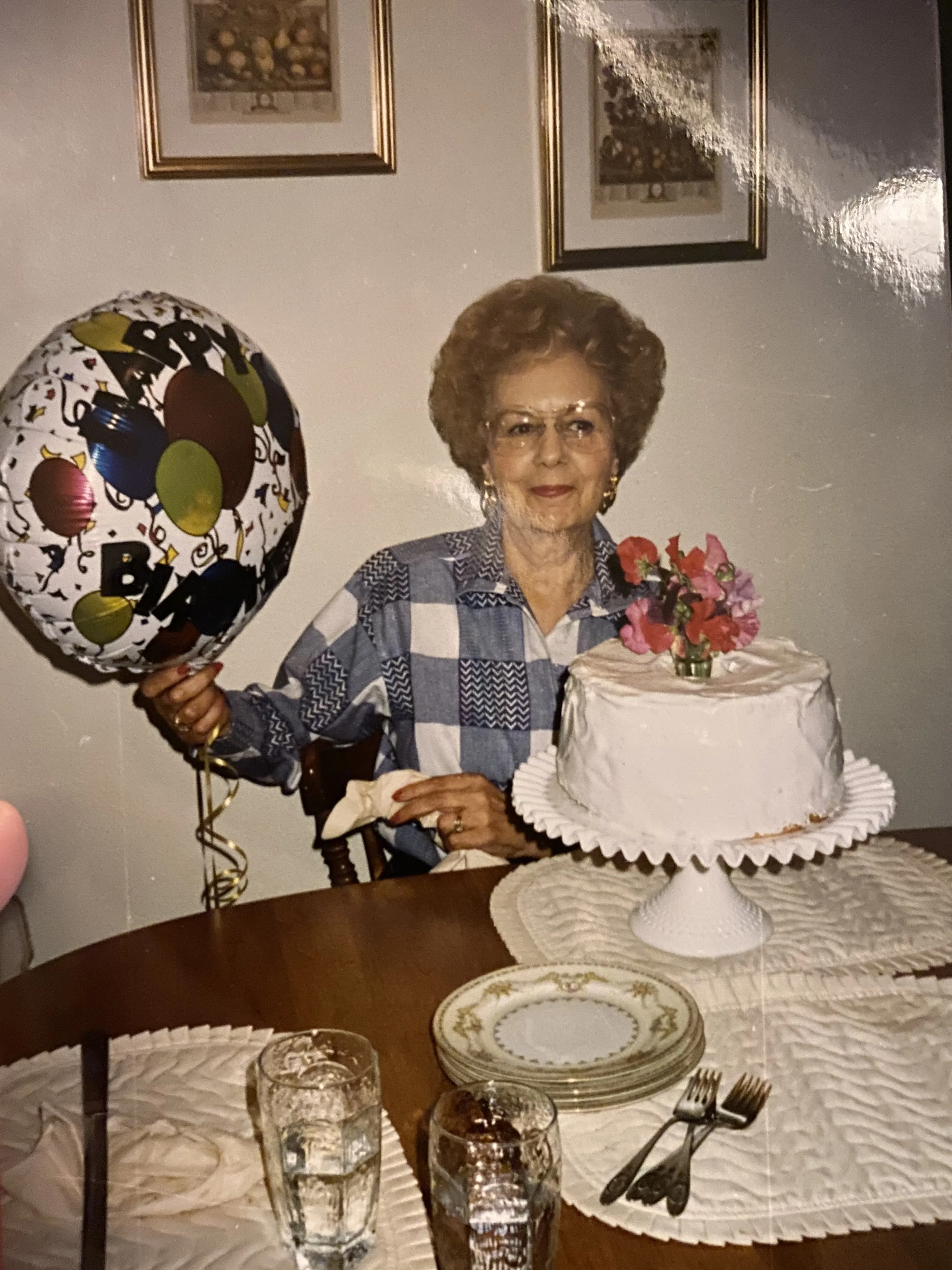 My grandma Idyll at her birthday, holding a Happy Birthday balloon next to a white frosted cake topped with flowers.