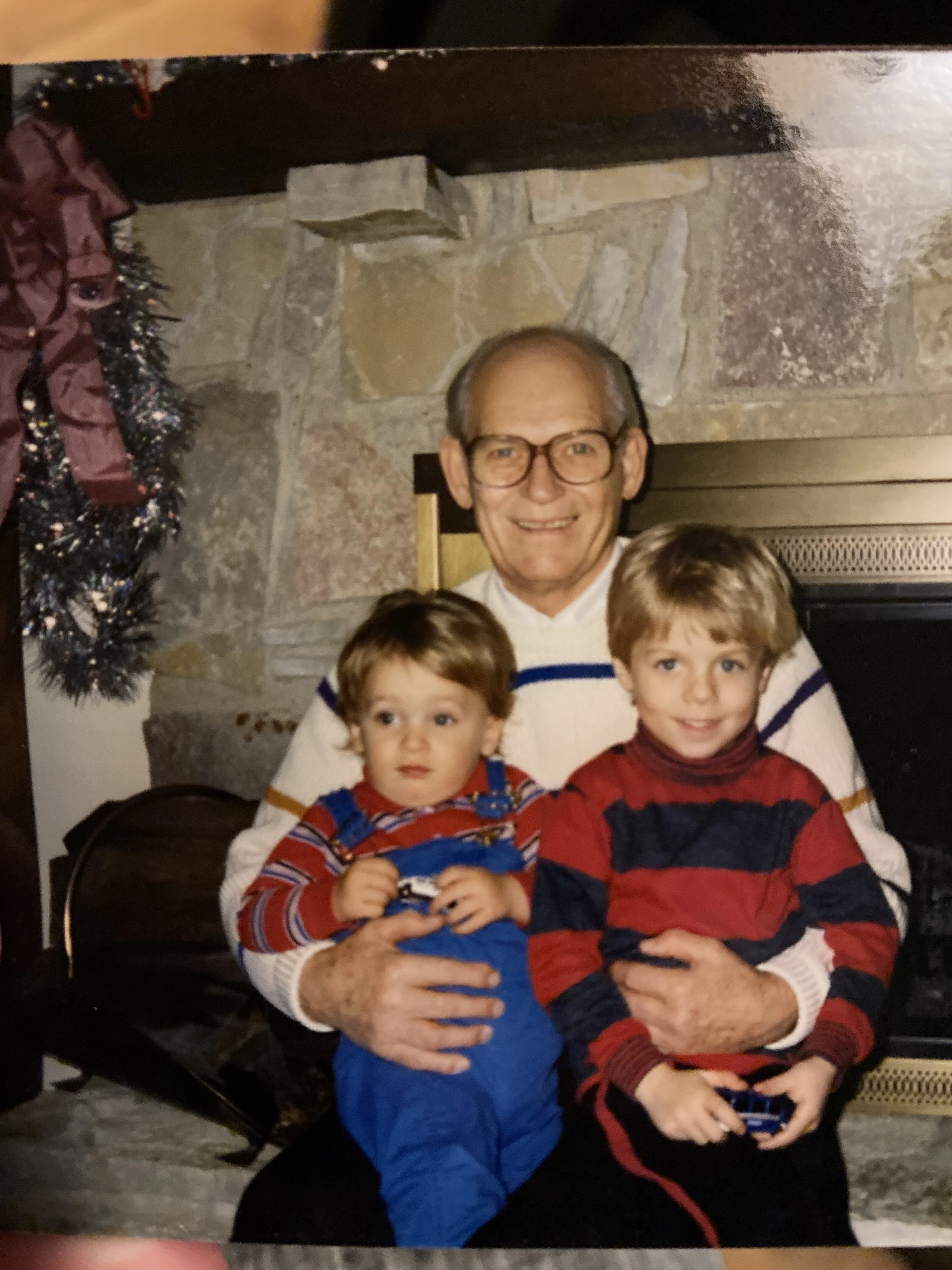My grandpa Czar in glasses and a striped sweater, sitting by a stone fireplace at Christmas, holding me as a toddler and my older brother on his lap.