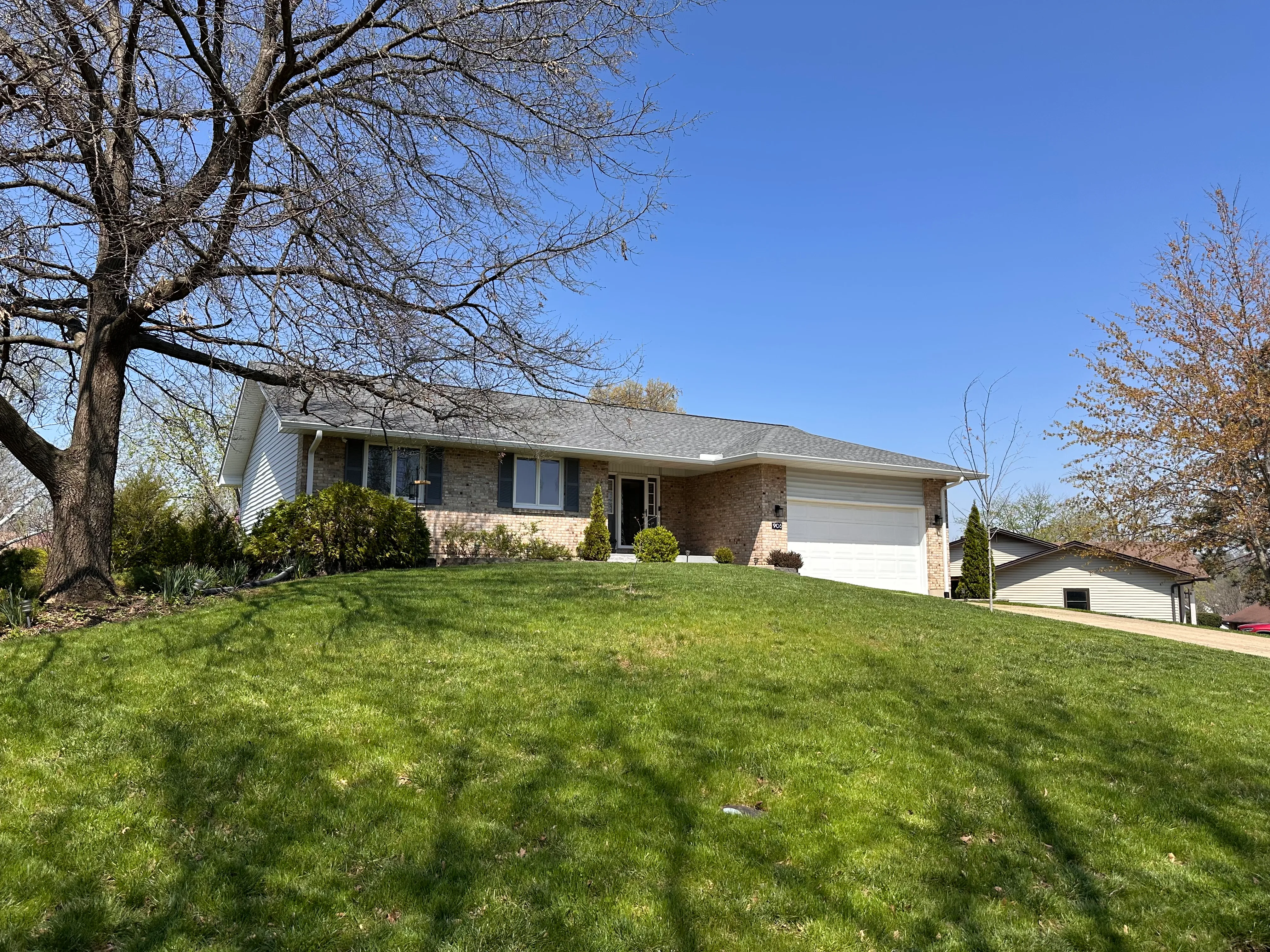 The same house in 2026, photographed from the same angle, the two large front-yard trees now gone and replaced by smaller ones, with a clearer view of the sky and a neighbor's outbuilding to the right.