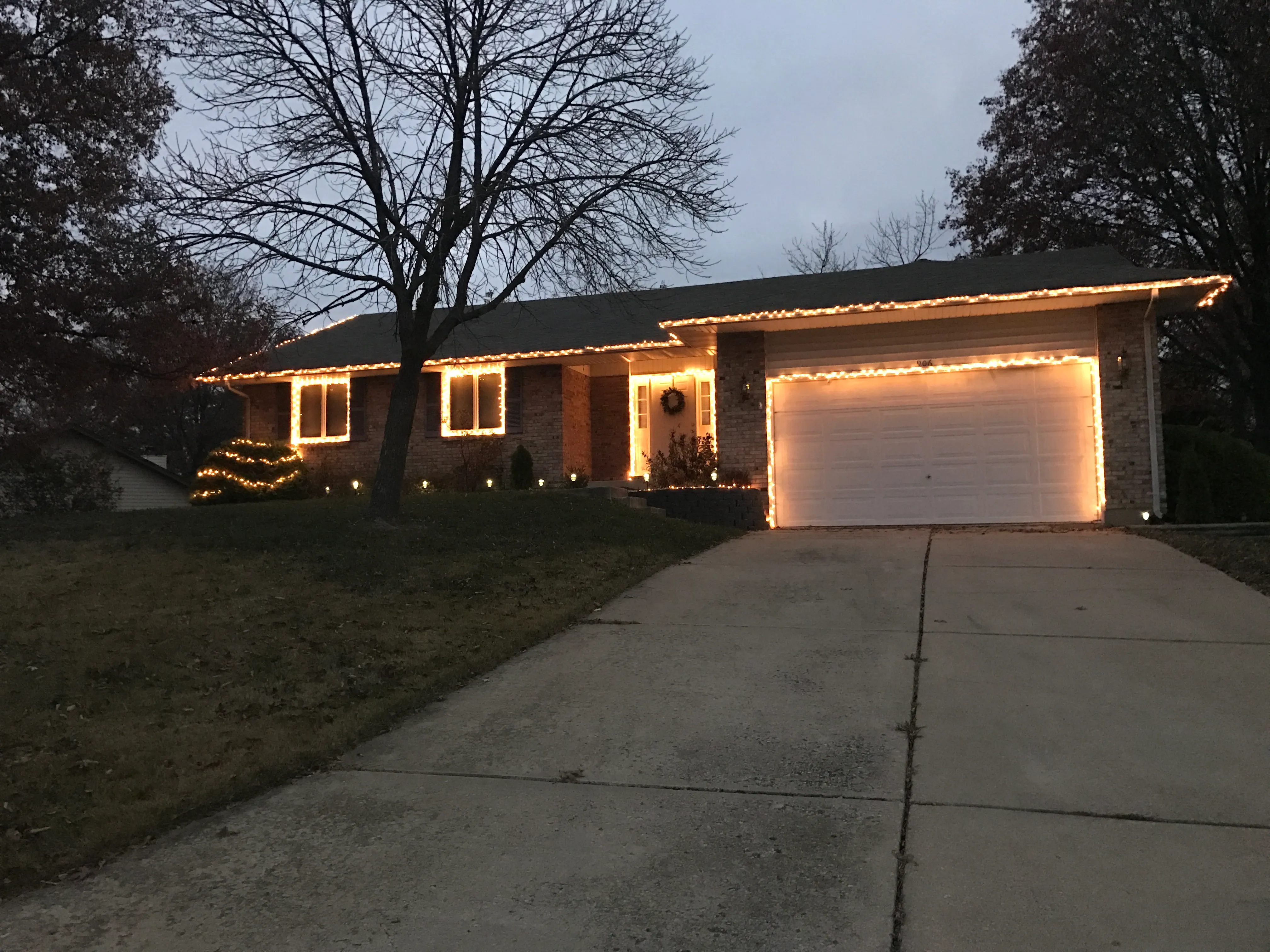 The house lit up with white Christmas lights along the roofline and bushes at dusk, snow-dusted lawn out front.