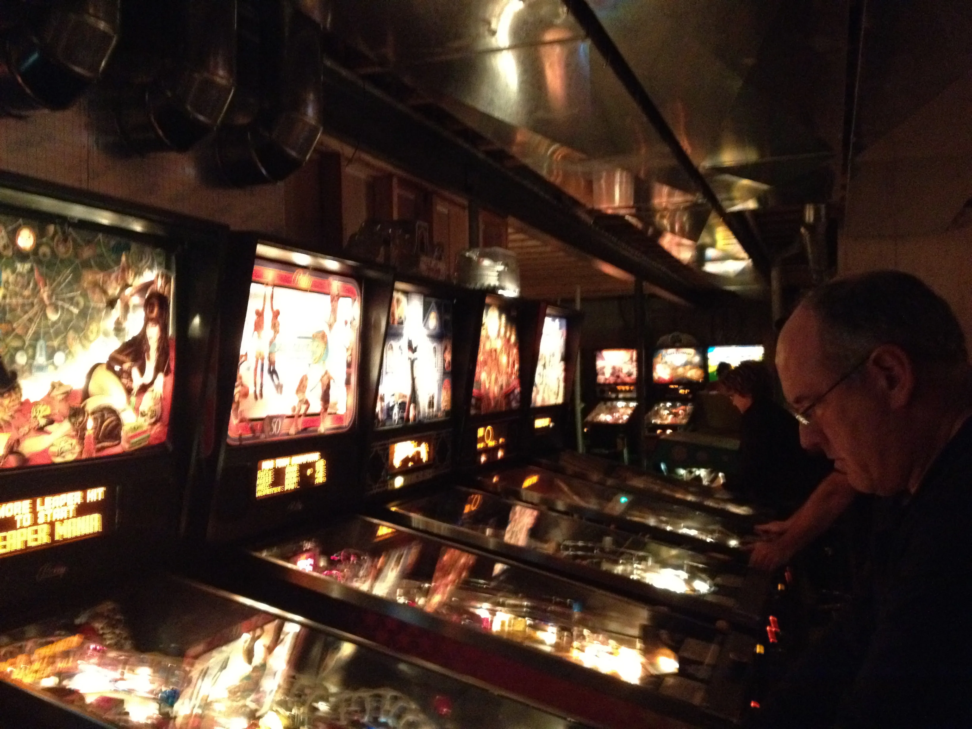 A dark row of glowing pinball machines, someone mid-game at the far end
