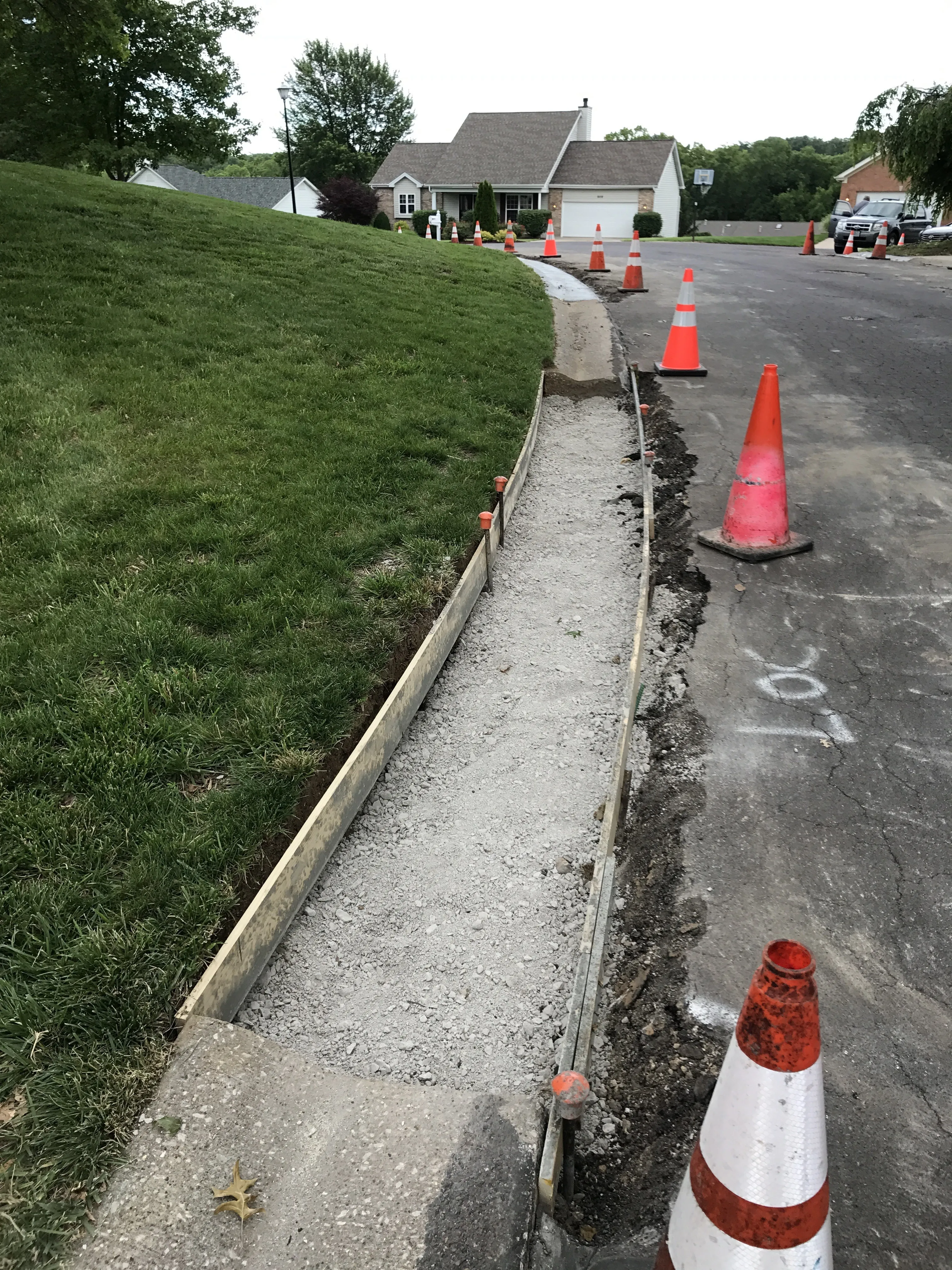 The street in front of the house torn up mid-replacement, a freshly poured concrete gutter running along the curb, orange traffic cones, and neighbors' houses in the background.