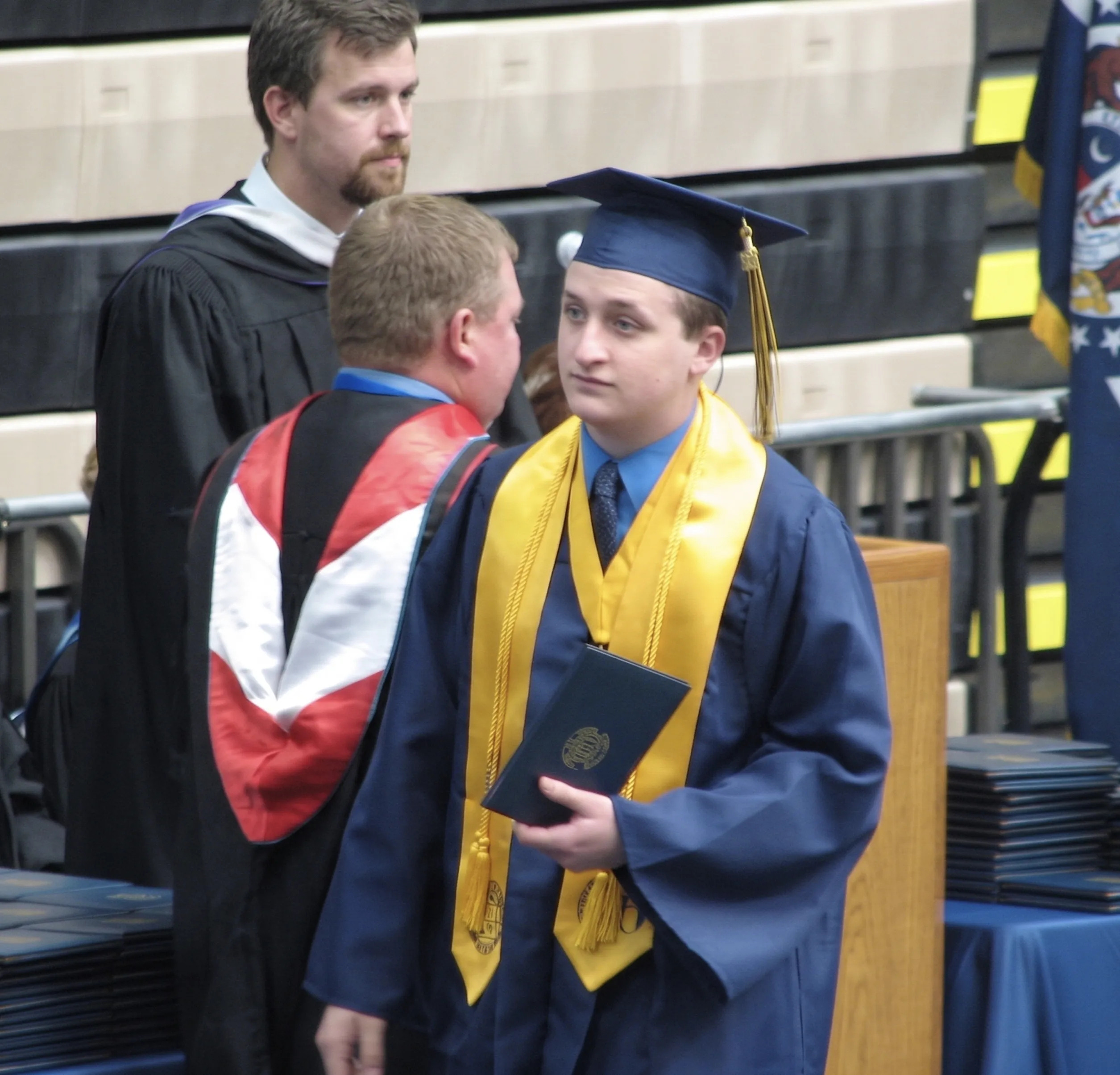 Me in a navy cap and gown with a gold honor stole, holding my diploma, on stage at my Holt High School graduation.