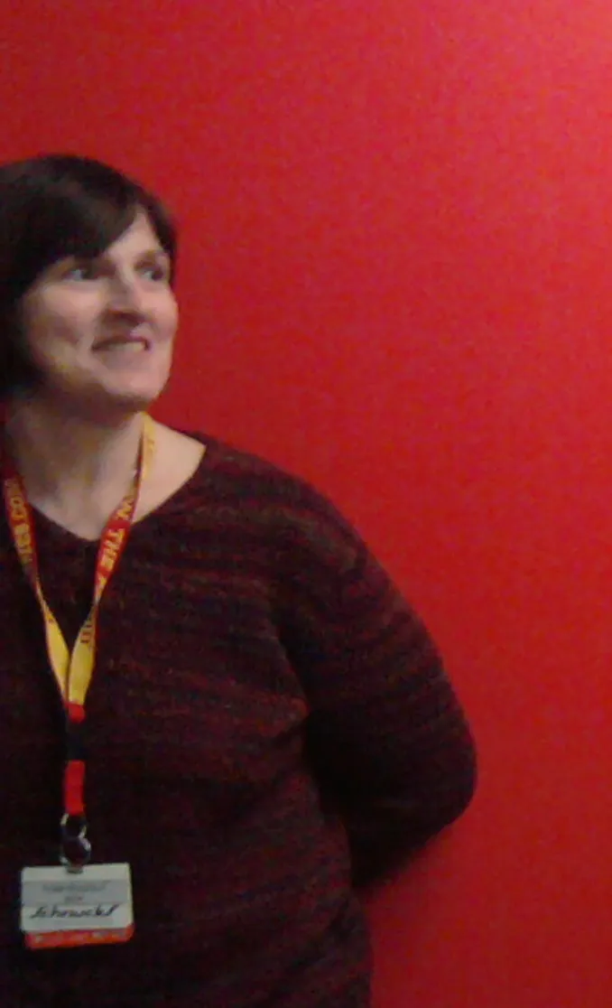 Toni at a work gathering, smiling and looking off to the side, wearing a maroon sweater and a work lanyard, against a red wall.