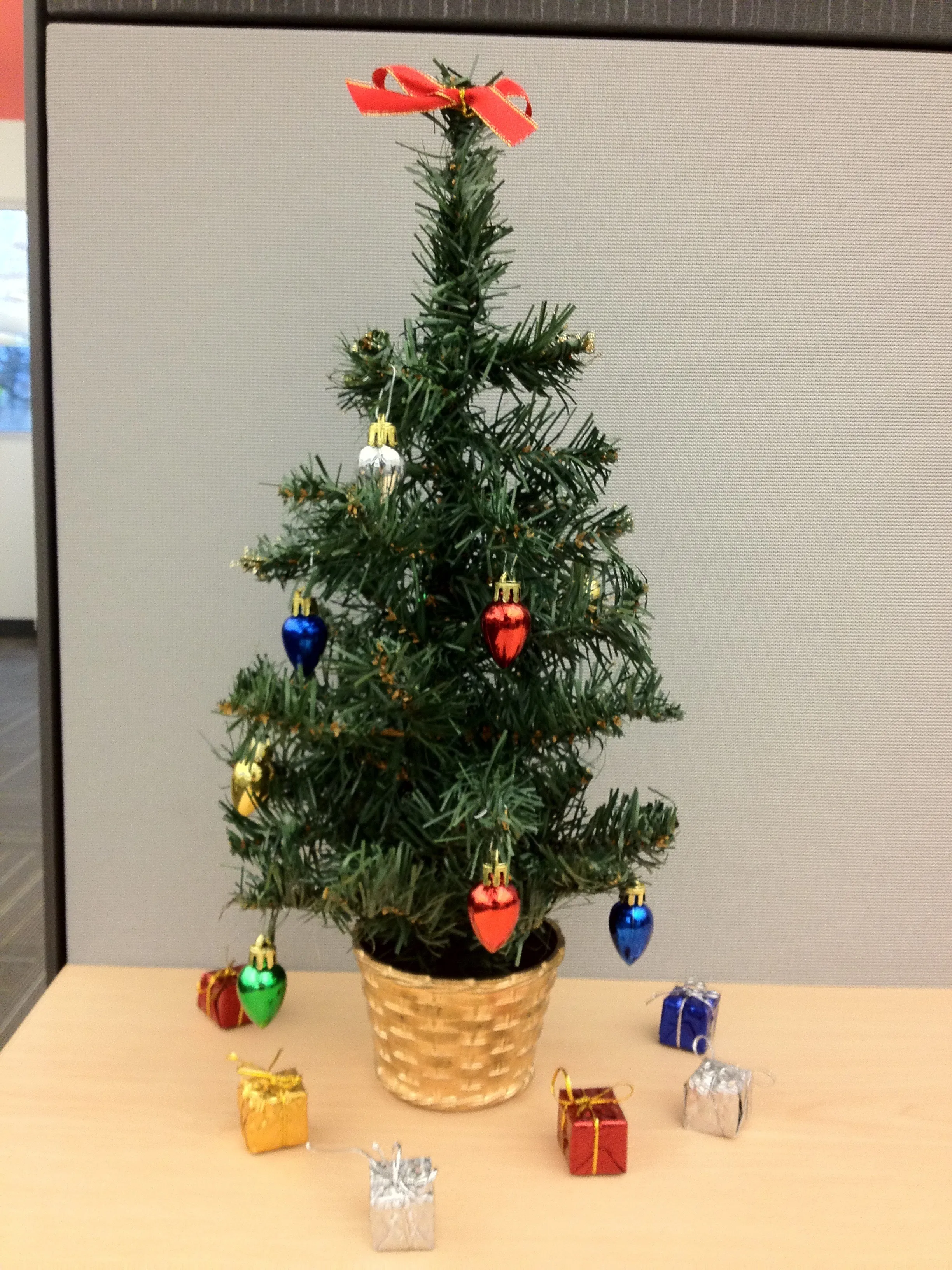 A small artificial Christmas tree on an office desk, decorated with a red bow and bulb-shaped ornaments, sitting in a gold basket with tiny wrapped presents around the base.