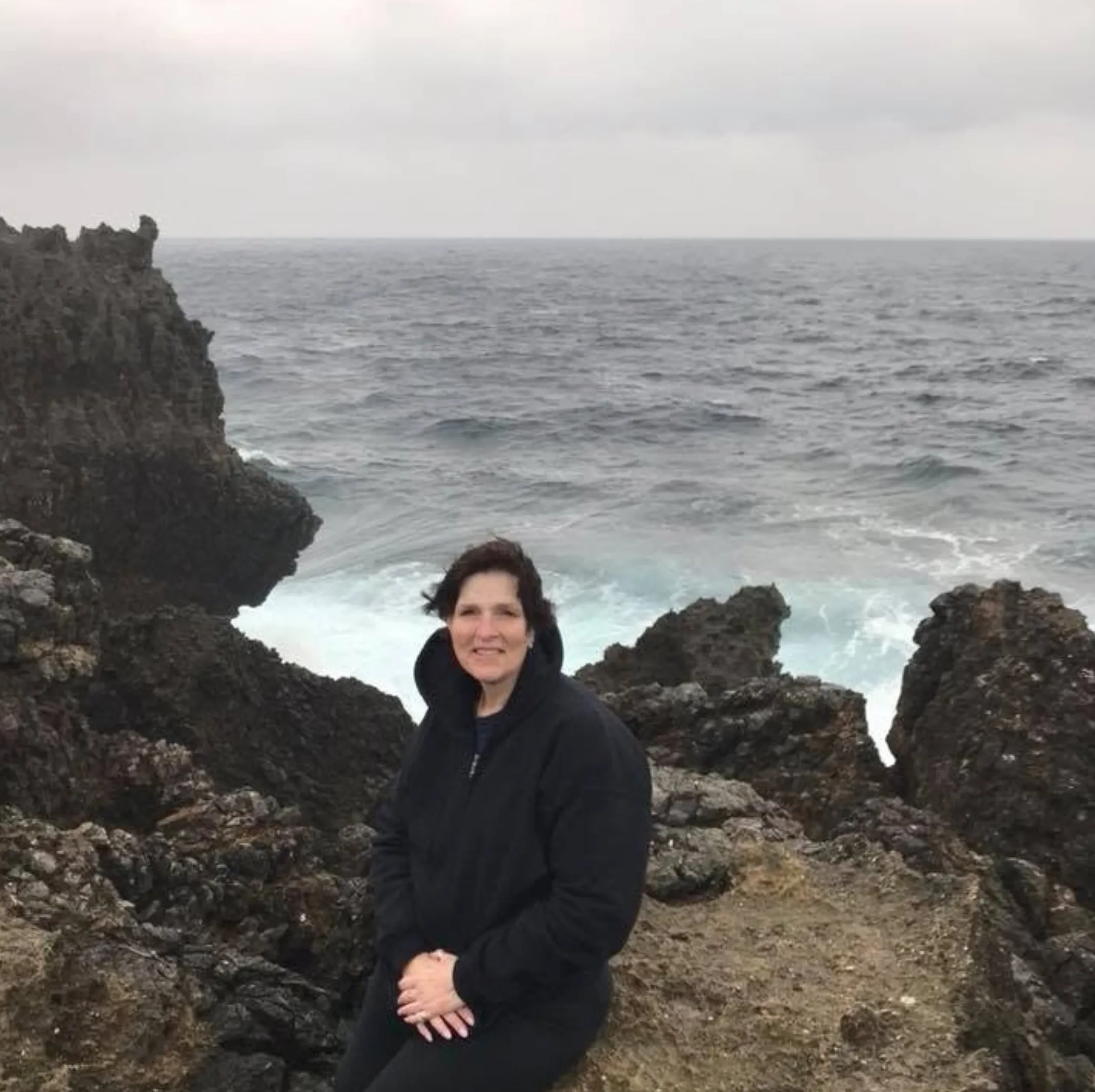 Toni sitting on dark volcanic rocks at the edge of a gray, choppy ocean, smiling, in a black fleece jacket.